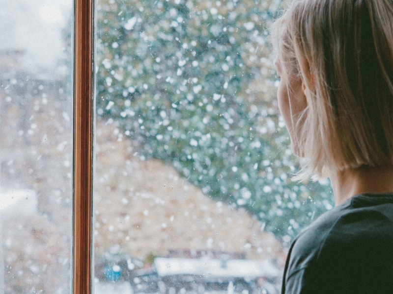 Woman standing by a window watching snow fall, appearing thoughtful and sad, suggesting grief during the holiday season.