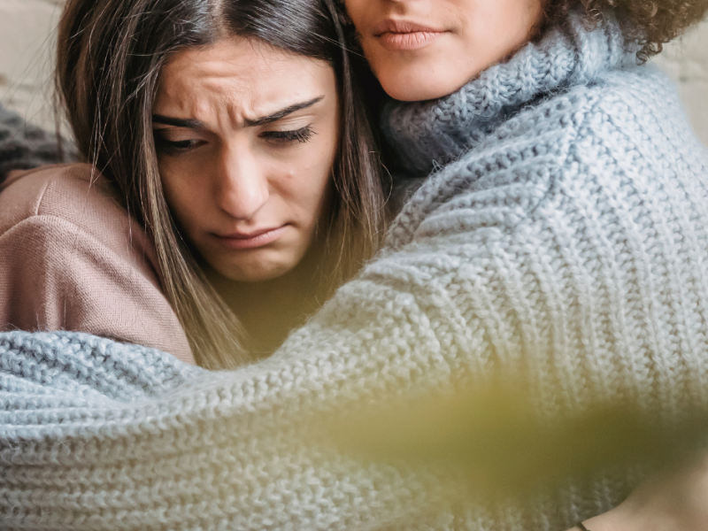 A distressed woman is comforted by a supportive friend, who holds her in a gentle embrace. The woman’s expression shows emotional pain, while the comforting presence highlights the importance of compassionate support after an abortion.