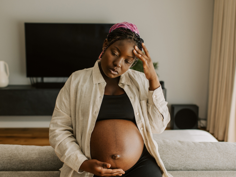 A visibly pregnant woman sits on a couch, resting one hand on her belly and the other on her forehead, appearing overwhelmed and deep in thought. Her expression suggests emotional strain, highlighting a need for support during pregnancy.