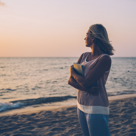 Woman standing on a beach at sunset, holding a journal and looking out at the ocean, symbolizing emotional healing and reflection after abortion.